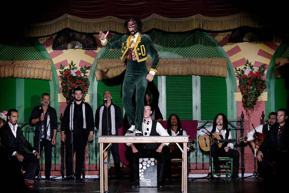 Performer dances on a table onstage, surrounded by musicians and singers.