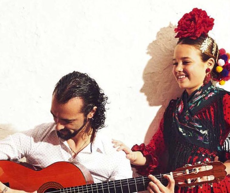 Man playing guitar beside smiling woman in traditional attire against white wall.