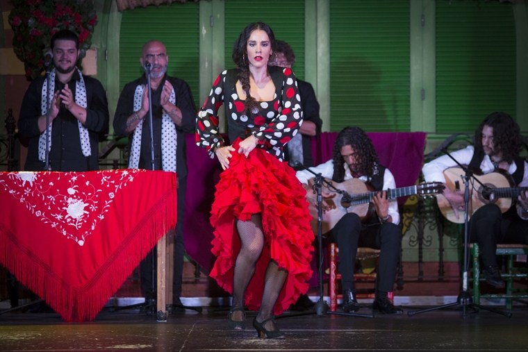 Flamenco dancer in red dress performs on stage with musicians and clapping audience.