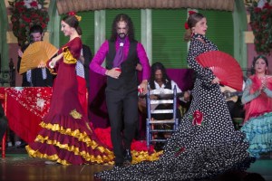 Flamenco dancers in vibrant costumes with fans on stage, musicians in background.
