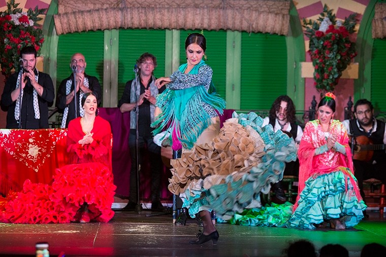 Flamenco dancer in colorful dress performs on stage with musicians clapping.