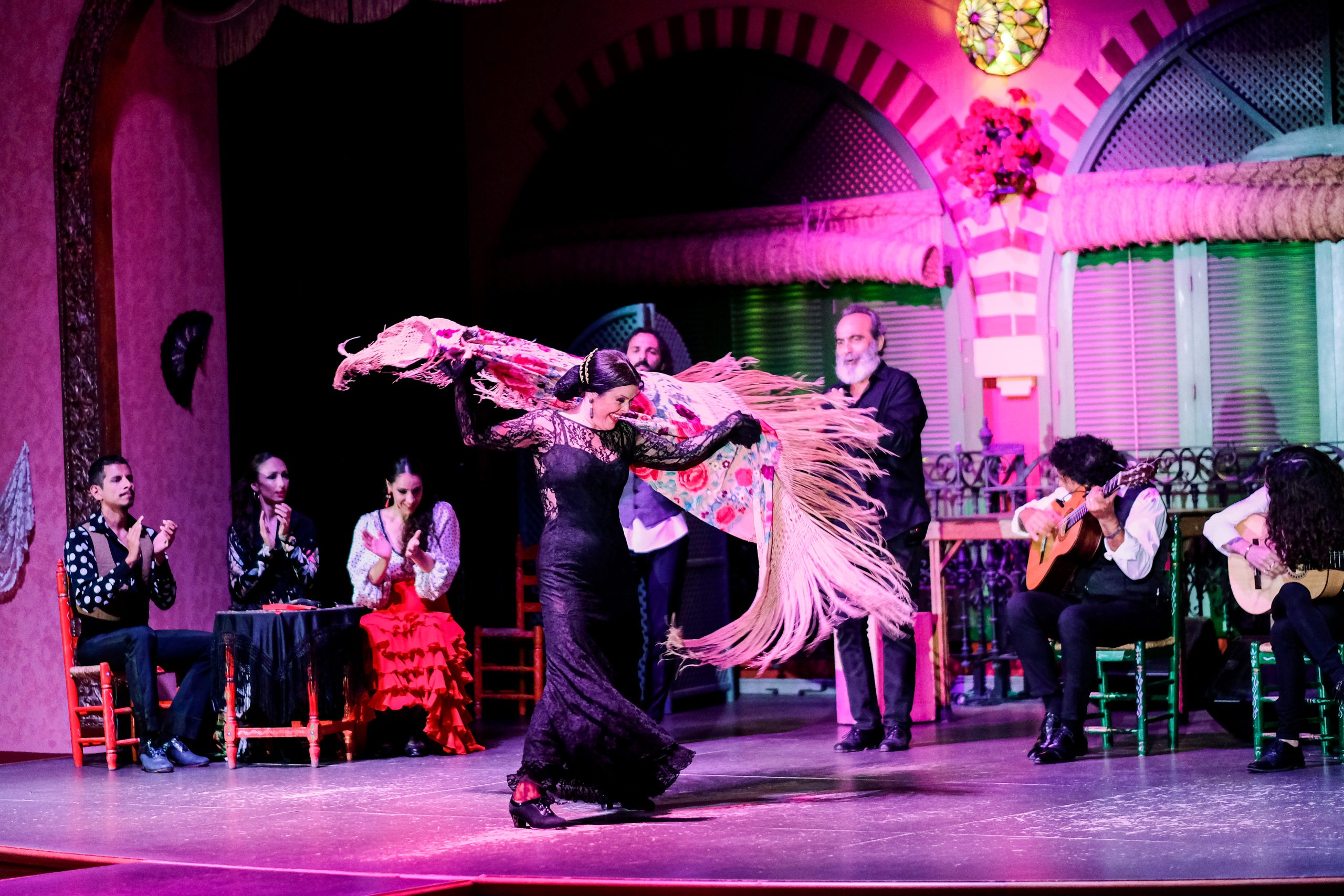 Flamenco dancer in vibrant dress twirls shawl, musicians and audience in background.