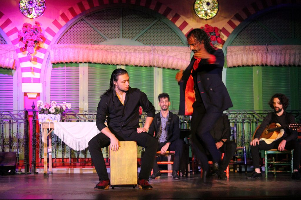Flamenco performance with dancer, guitarist, and percussionist on a colorful stage.