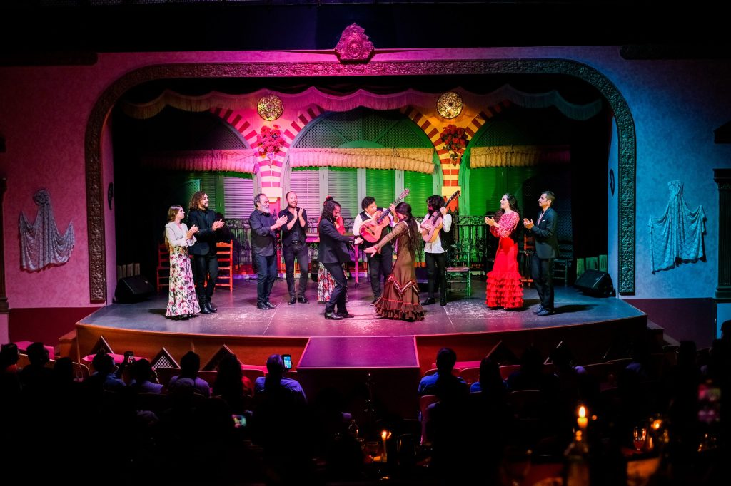 Group of performers on stage in colorful costumes, with audience watching.