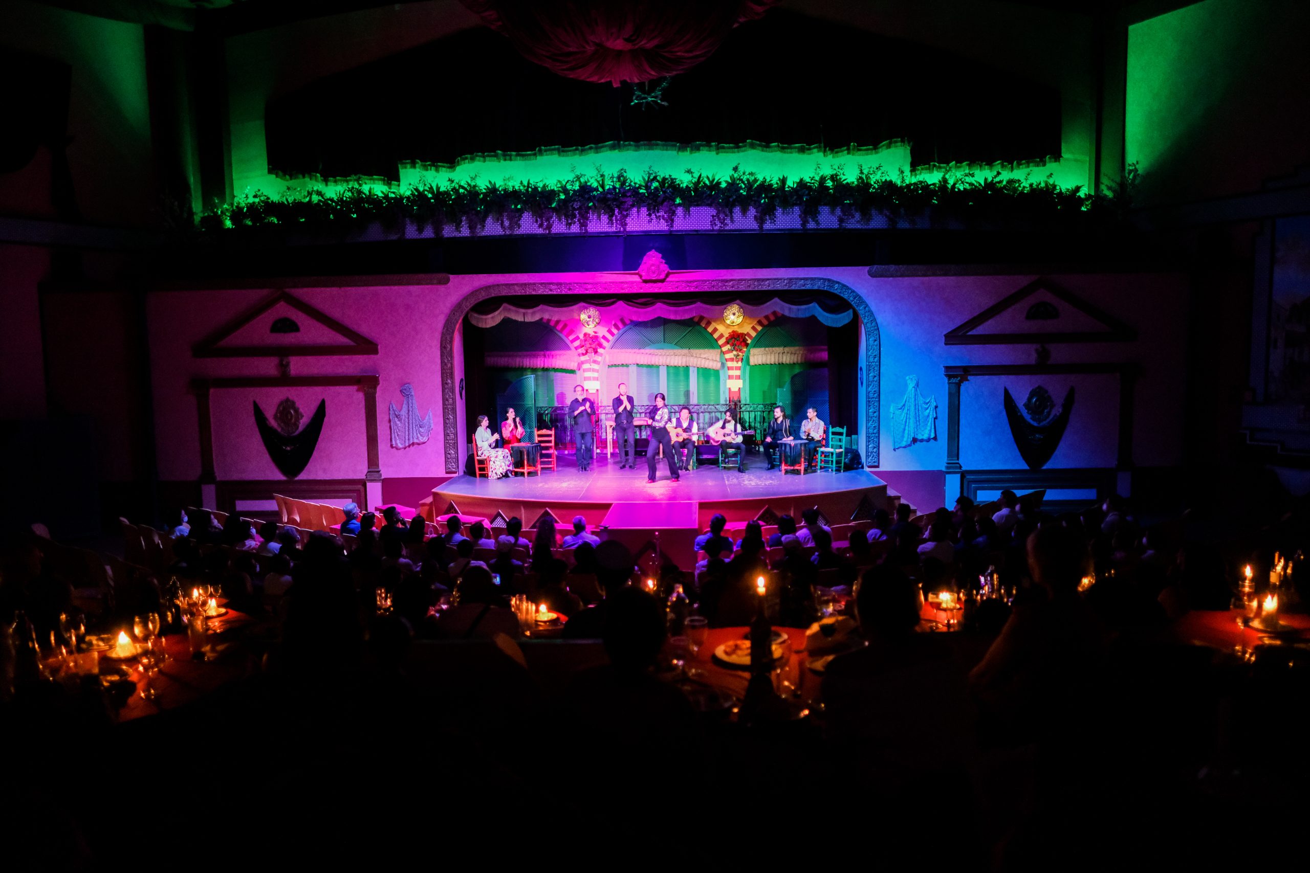 Dimly lit stage with performers and seated audience in a colorful theater.