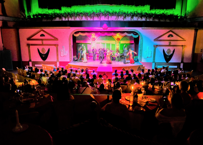 People watch a vibrant stage performance with colorful lighting in a theater.