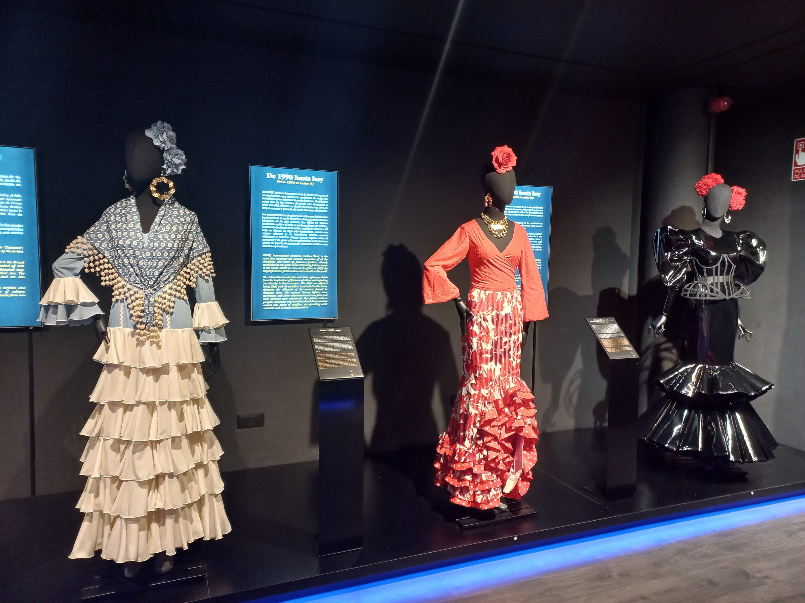 Three mannequins display colorful flamenco dresses with ruffles in a dimly lit exhibit.