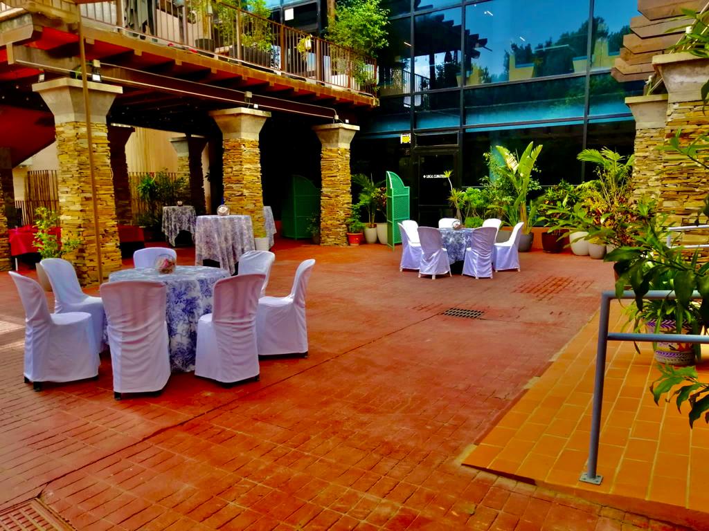 Outdoor venue with round tables, white chairs, stone columns, and potted plants.