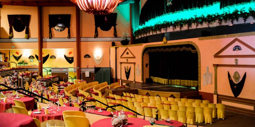 Vintage theater with yellow seats, tables set for dining, and a stage with ornate decorations.