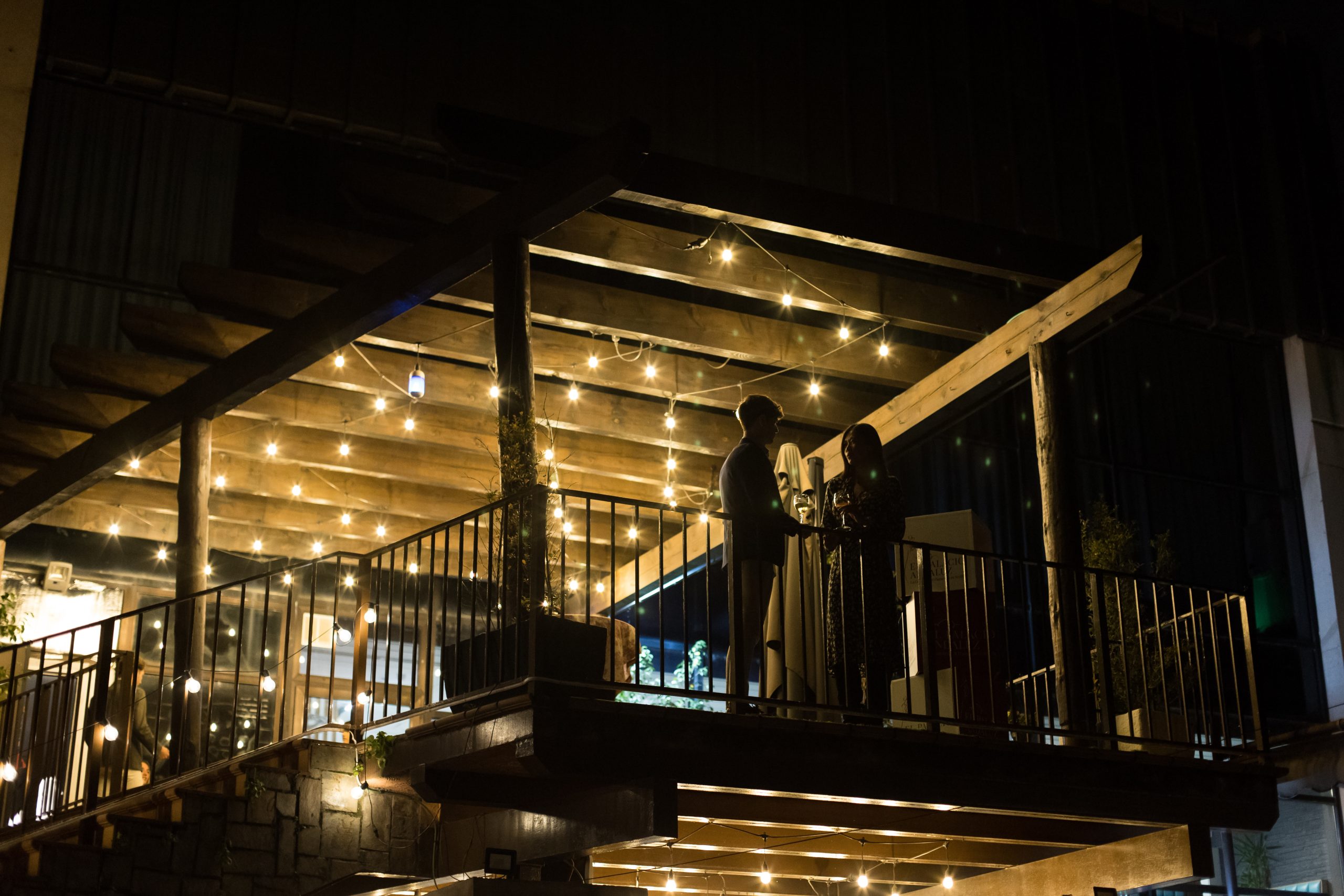 Two people standing on a dimly lit balcony with string lights at night.