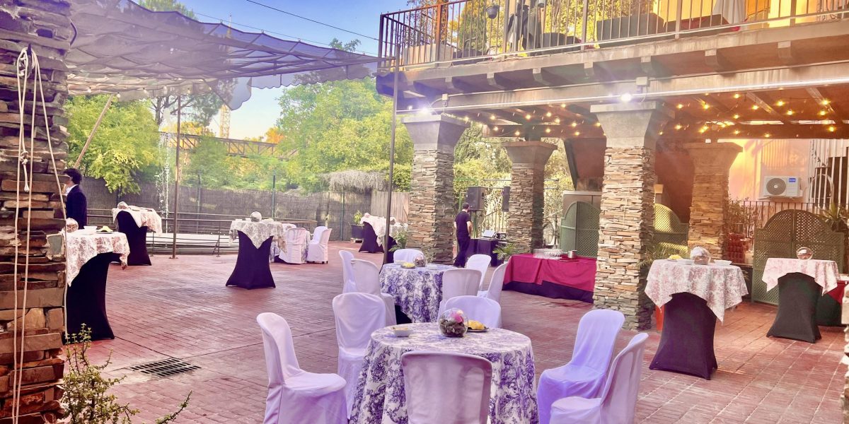 Outdoor restaurant with tables covered in white and black cloths, surrounded by stone columns and plants.
