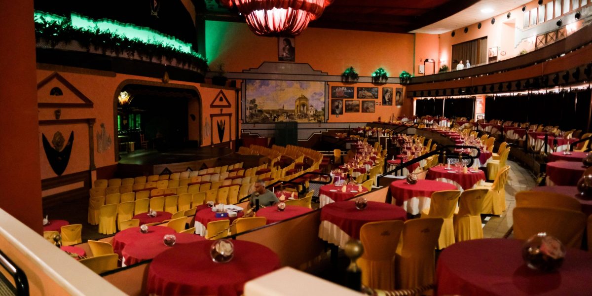 Vintage theater with red tables and yellow chairs, stage, wall paintings, and dim lighting.