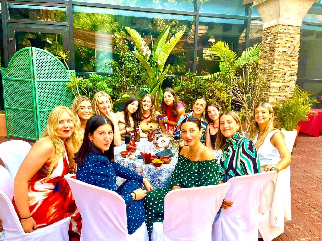 Group of women seated around a table outdoors, smiling, with plants in the background.