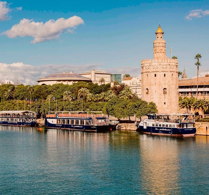 Historic tower beside a river with moored boats and blue sky.