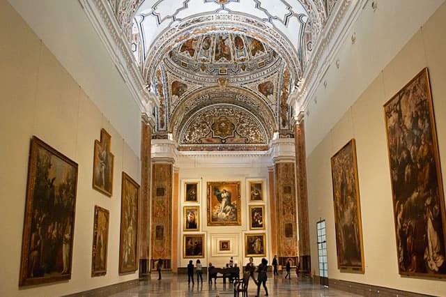 Ornate museum hall with large paintings and vaulted, decorative ceiling.