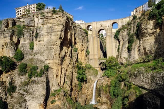 Stone bridge spanning a deep gorge with waterfall and cliffside buildings.