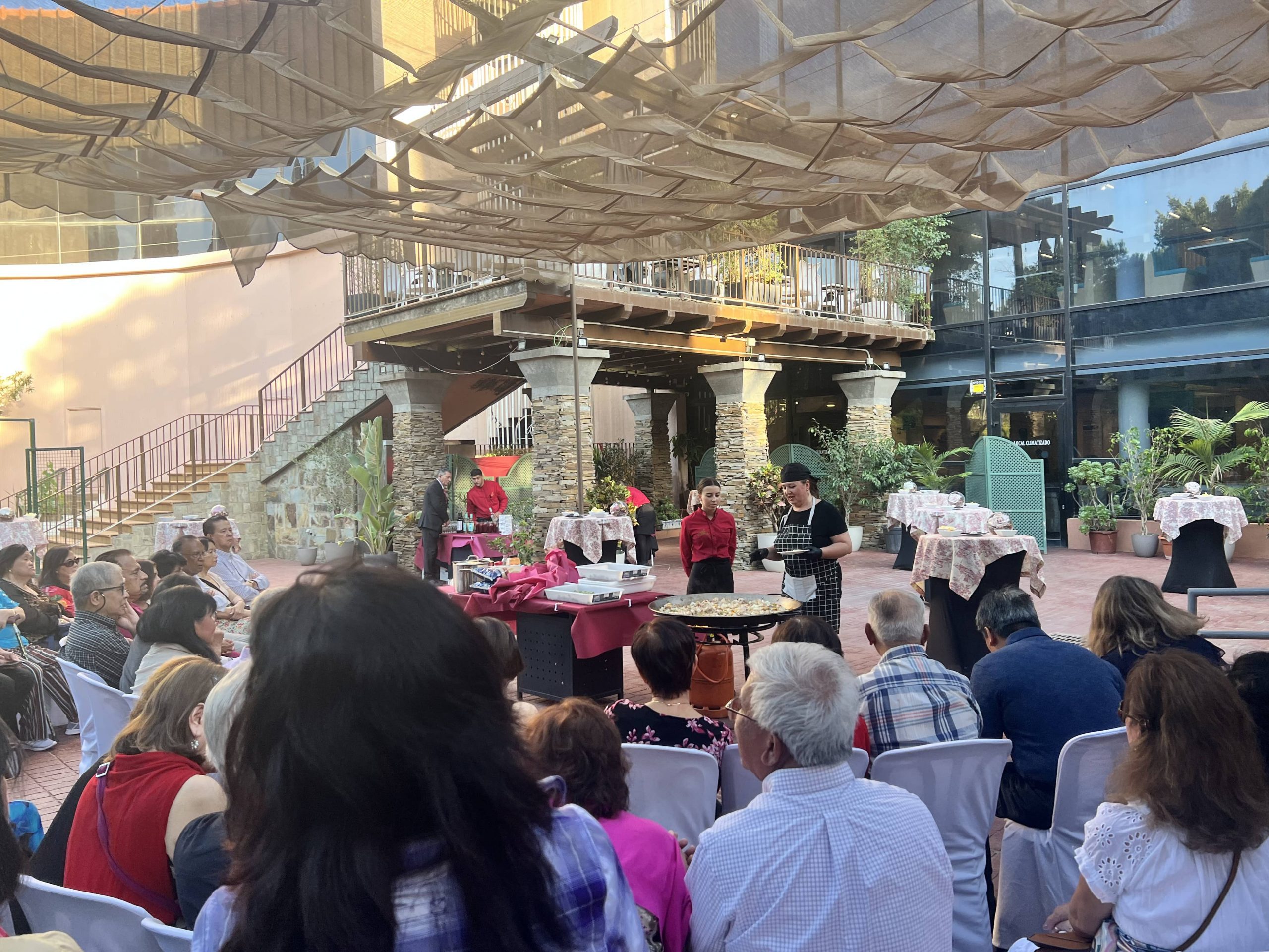Crowd watching cooking demonstration in outdoor courtyard under canopy.
