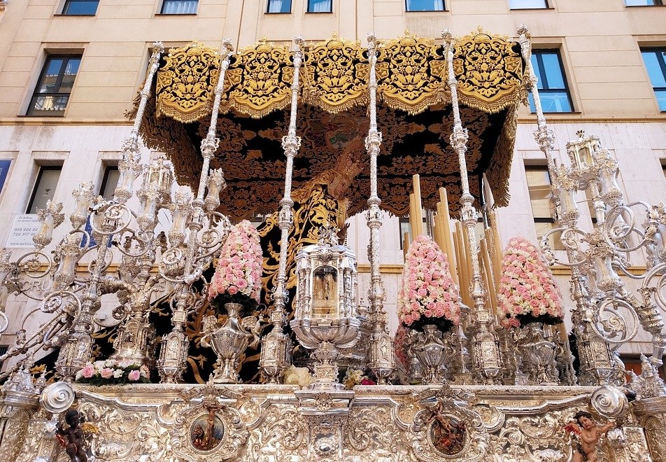 Ornate religious float with silver details and pink flowers in a procession.