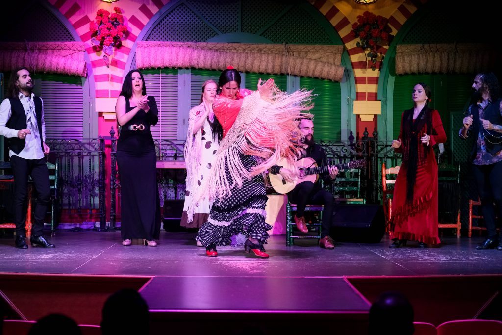 Flamenco performers on stage with a dancer in a red top and shawl, surrounded by guitarists and singers.