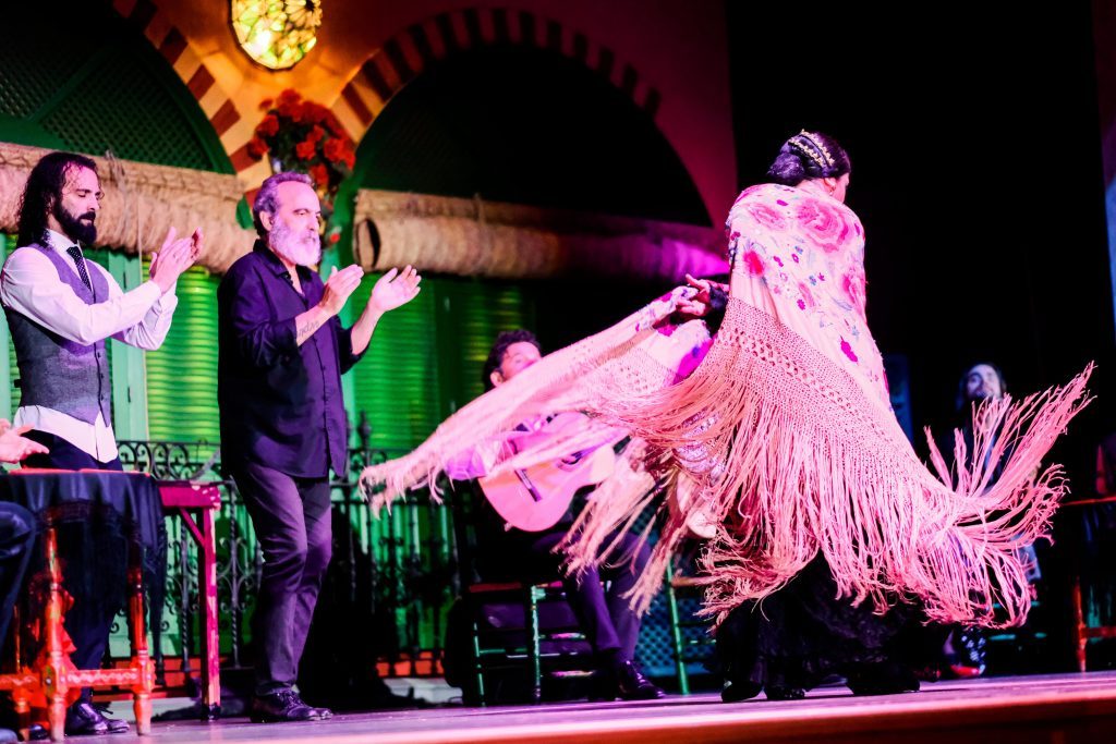 Dancer with shawl performing flamenco on vibrant stage with musicians.
