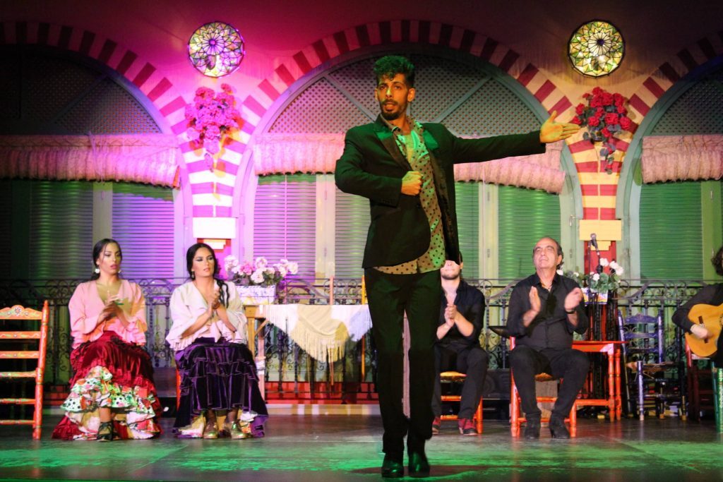 Flamenco dancer performs on stage with musicians seated in colorful, decorated setting.