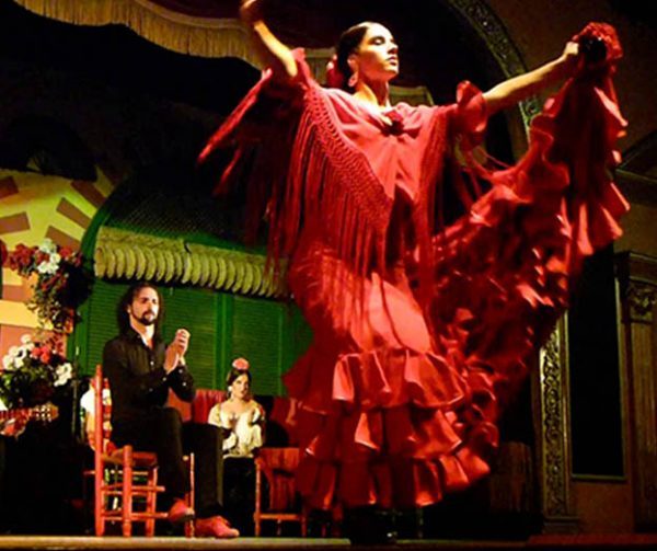 Flamenco dancer in red dress performing on stage with musicians in the background.
