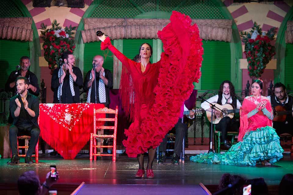 Flamenco dancer in red dress performs on stage with musicians and audience.