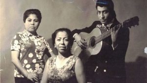 Three people; two women seated, one standing with a guitar.