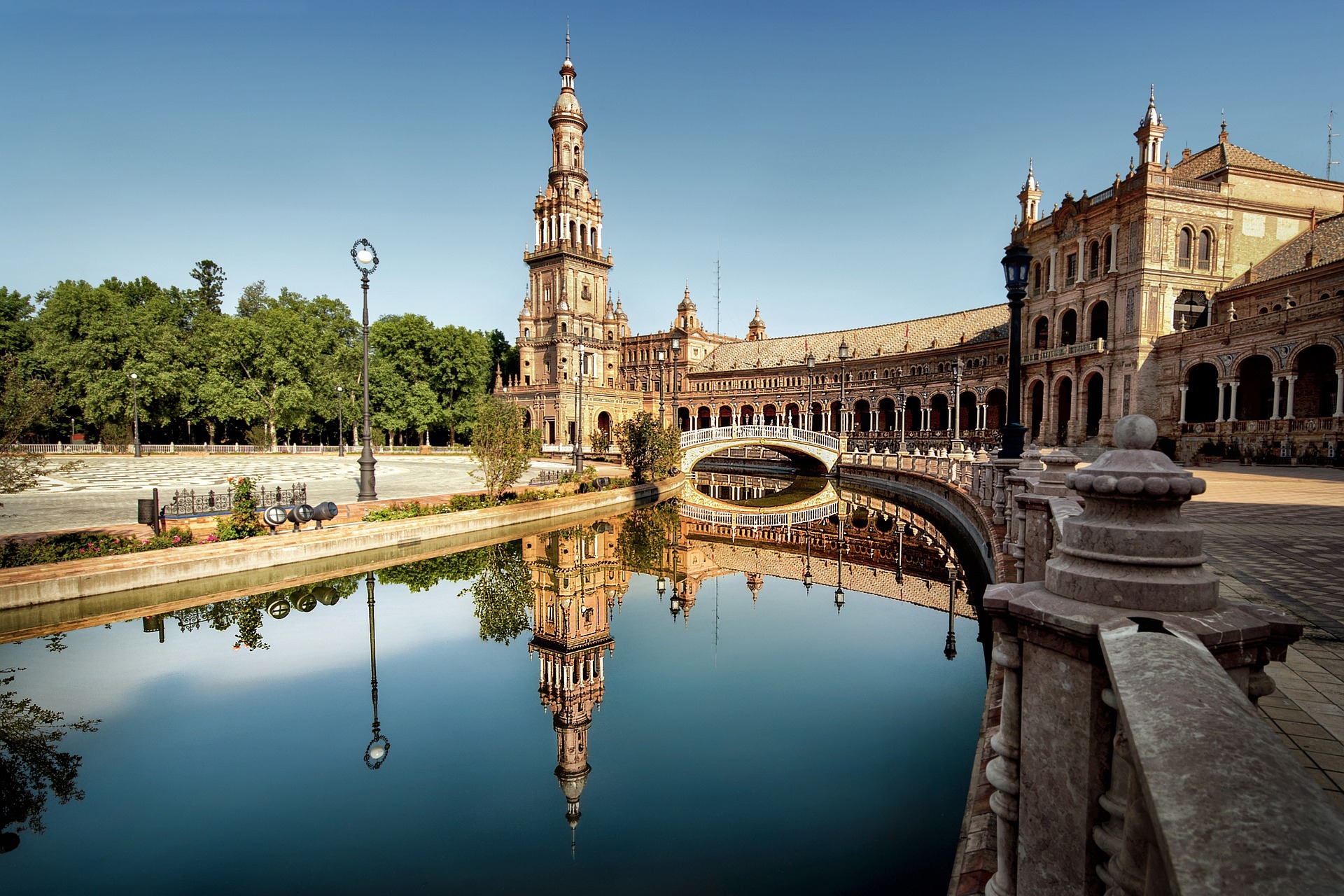 Scenic view of plaza with large building, tower, and arched bridge reflecting in a calm body of water.