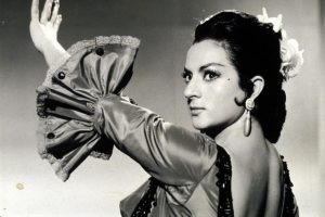 Woman in traditional dress posing with raised arm, wearing hoop earrings and flowers in her hair.