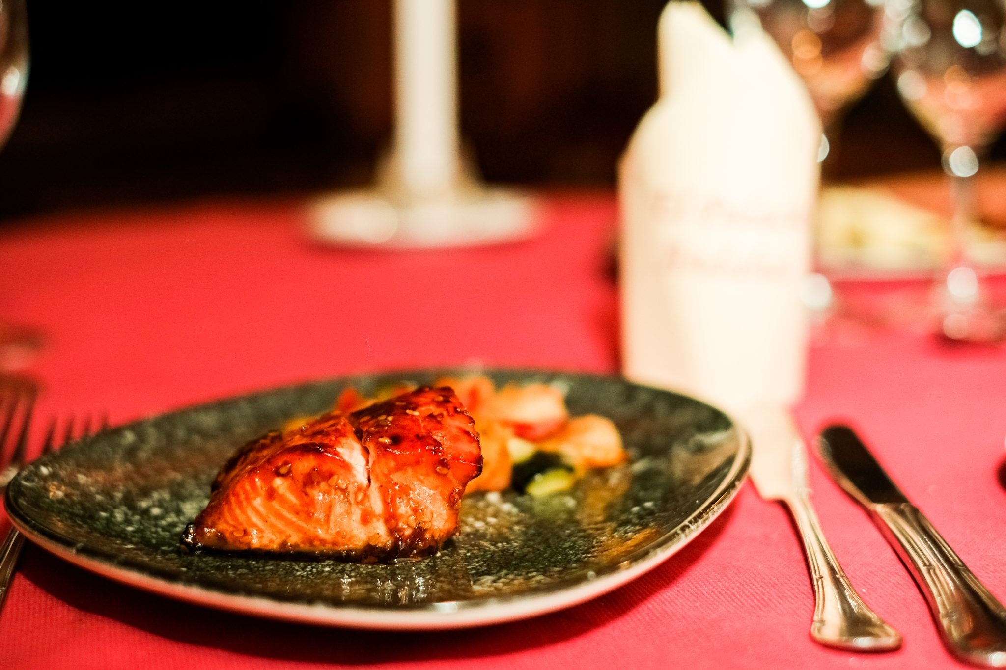 Grilled salmon on a dark plate with blurred cutlery on red tablecloth.